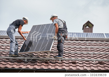 Men technicians lifting photovoltaic solar modul on roof of house. Electricians in helmets mounting solar panel system outdoors. Concept of alternative and renewable energy. 101639639