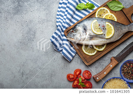 Raw fish dorado with ingredients lemon, fresh basil, cut cherry tomatoes, uncooked rice on wooden cutting board with knife on rustic stone table top view, cooking healthy fish dorado. Copy space Raw fish dorado with ingredients lemon, fresh basil, cut cherry tomatoes, uncooked rice on wooden cutting board with knife on rustic stone table top view, cooking healthy fish dorado. Copy space 101641305