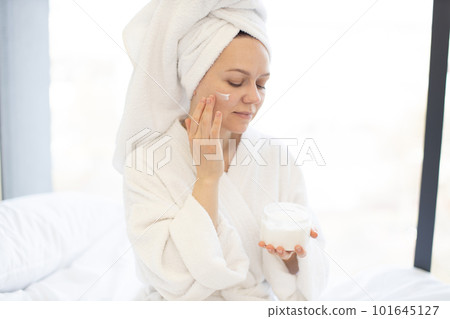 Woman applying facial cream from jar in bedroom at home 101645127