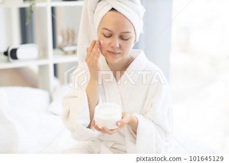 Woman applying facial cream from jar in bedroom at home 101645129