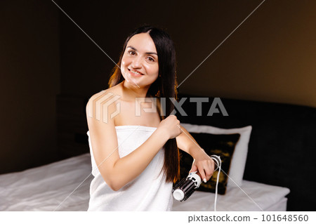 A beautiful woman in bath towel standing in room dries and styles her hair with a hair dryer comb. Morning routine. A beautiful woman in bath towel standing in room dries and styles her hair with a hair dryer comb. Morning routine. 101648560