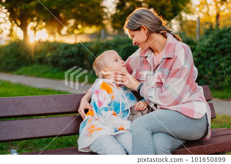 mother and son sit on a park bench in the rays of the setting sun. the concept of a family. Mother's Day. beautiful girl (mother) with a boy (son) in the park in the park are sitting on a bench at mother and son sit on a park bench in the rays of the setting sun. the concept of a family. Mother's Day. beautiful girl (mother) with a boy (son) in the park in the park are sitting on a bench at 101650899