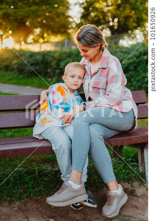 mother and son sit on a park bench in the rays of the setting sun. the concept of a family. Mother's Day. beautiful girl (mother) with a boy (son) in the park in the park are sitting on a bench at 101650926