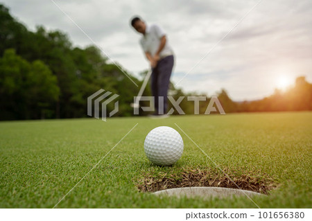 White golf ball rolling down golf hole on putting green with evening golf course. 101656380