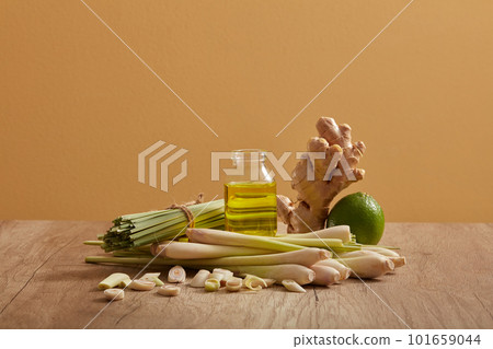 A glass transparent jar of essential oil extracted from lemongrass, ginger and lemon is displayed between bunch of lemongrass, a lemon and a ginger on wooden table 101659044