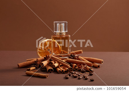 A glass transparent bottle filled with perfume extracted from coffee beans, cinnamon sticks and dried orange slice, displayed on dark minimalist background. Product mockup 101659054