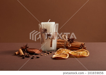 A white candle putted on a transparent glass and a bunch of cinnamon sticks, coffee beans, star anise and dried orange slices decorated around. 101659056