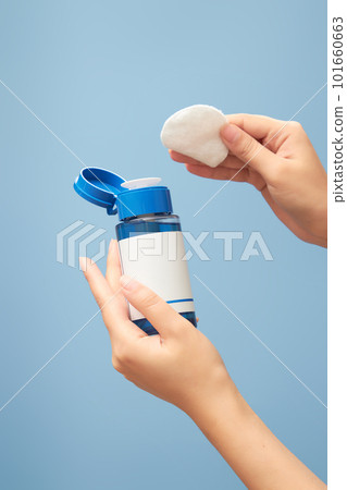 Front view of the other hand holding an empty bottle mockup makeup remover bottle and the other hand holding a cotton pad on blue background. Empty space for label design. 101660663