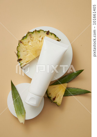 A white tube placed on round shaped podiums, displayed with pineapple leaves and slices. Empty label to promote beauty product extracted from pineapple (Ananas comosus) 101661405