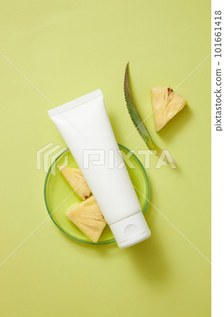 Top view of an empty label white tube placed on a glass petri dish containing essence extracted from pineapple and pineapple slices. Cosmetic product mockup 101661418