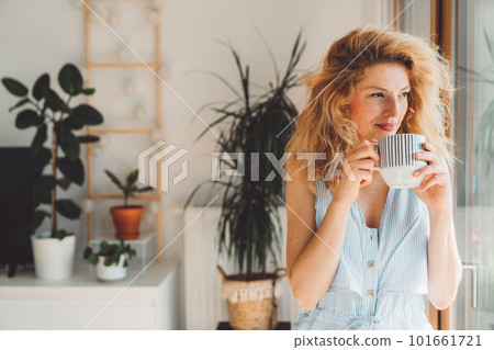 Waist up portrait woman with blonde curly hair holding a cup of tea standing by the window looking out, plants in the background Waist up portrait woman with blonde curly hair holding a cup of tea standing by the window looking out, plants in the background 101661721