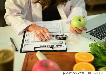 Cropped image of nutritionist holding an apple and prescribing recipe at desk with fresh fruit. Healthcare and diet concept 101670435