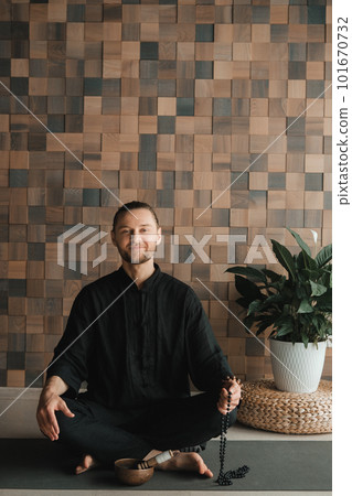 Portrait of a young man in a black kimano sitting in a lotus position on a gym mat in the interior 101670732
