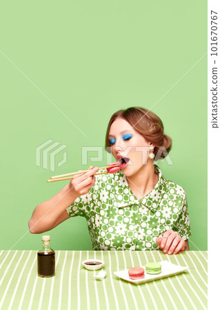 Pretty, stylish, young girl eating sweet macarons with chopsticks against green studio background. Soy sauce taste 101670767