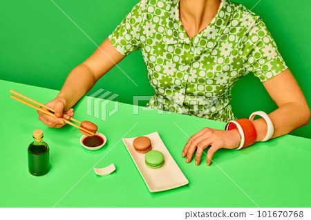 Cropped image of girl putting sweet macarons into soy sauce with chopsticks against green studio background. Extraordinary taste combination 101670768