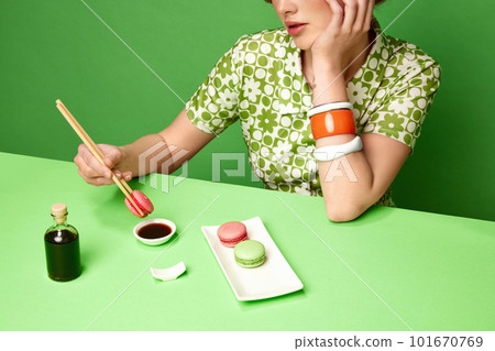 Cropped image of girl putting sweet macarons into soy sauce with chopsticks against green studio background. 101670769