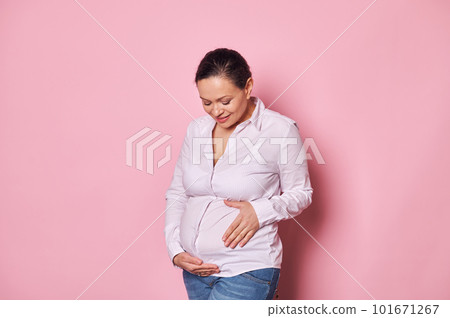 Delightful happy middle-aged pregnant woman, smiling cutely while touching and caressing her belly, enjoying her happy and carefree pregnancy, isolated on pink background. Happy Mother's Day concept 101671267