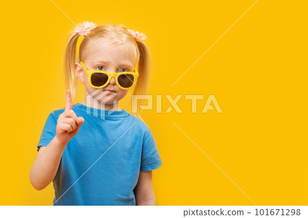 Portrait of little girl wears blue T-shirt and yellow sunglasses on yellow background. Child raised her index finger. Copy space, mock up Portrait of little girl wears blue T-shirt and yellow sunglasses on yellow background. Child raised her index finger. Copy space, mock up 101671298