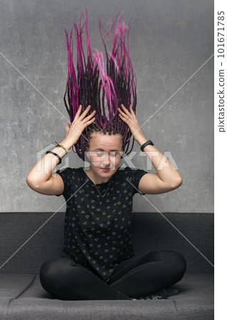 Portrait of young caucasian woman with long dreadlocks sitting on sofa. Girl waves her dreads. Vertical frame. 101671785