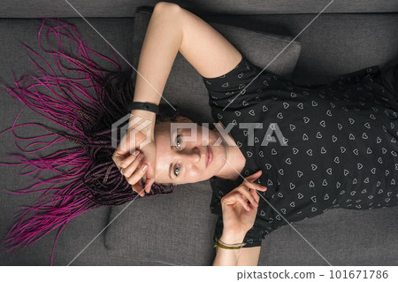 Portrait of pretty casually dressed caucasian young woman with dreadlocks looking into camera and lying on the sofa. 101671786