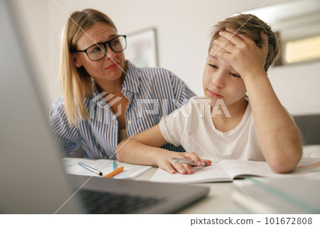 Tired sad kid sits at table with laptop and textbooks while mom help him with homework 101672808