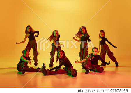 Art. Group of children, little girls in sportive casual style clothes dancing hip-hop, contemp dance against yellow studio background in neon light 101678489