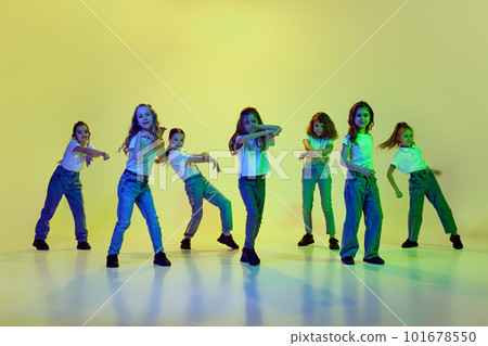 Dynamic image of little girls, children dancing against green studio background in neon light. Casual clothes. Hip-hop style 101678550