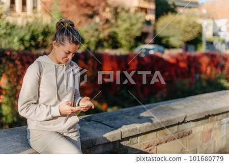 Young Fashionable Teenage Girl With Smartphone In Europian Park In Autumn Sitting At Smiling. Trendy Young Woman In Fall In Park Texting. Retouched, Vibrant Colors. Beautiful Blonde Teenage Girl 101680779