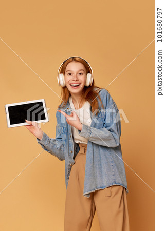 Portrait with student, girl wearing headphones holding tablet and pointing on empty screen with smile over ginger background. Online school 101680797