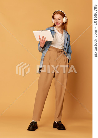 Portrait with student, girl wearing headphones holding tablet and looking on it over ginger background. Online lesson 101680799