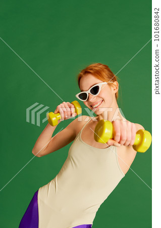 One young charming girl, woman wearing sunglasses doing workout with dumbbells and smiling at camera over green background. Upper body training One young charming girl, woman wearing sunglasses doing workout with dumbbells and smiling at camera over green background. Upper body training 101680842