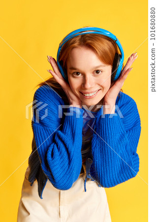 Portrait of young beautiful girl, teenager with red hair wearing headphones and smiling over yellow studio background. Happiness, joyful Portrait of young beautiful girl, teenager with red hair wearing headphones and smiling over yellow studio background. Happiness, joyful 101680880