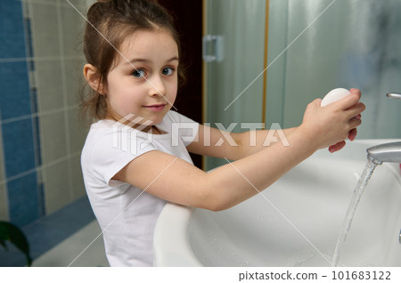 Beautiful little child girl smiling looking at camera, holding soap bar and washing her hands, standing by white ceramic sink at home bathroom. Cleanliness. Hygiene. Sanitary and health concept 101683122