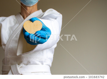 COVID-19 Pandemic Coronavirus concept. A young strong man in a white kimono for sambo, jiu jitsu and other martial arts with a blue medical gloves and medical mask. Wooden heart. 101683742