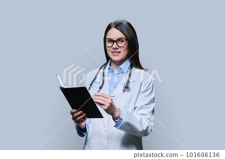 Young female doctor writes in notebook, on gray studio background. 101686136