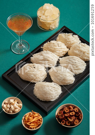 Ingredients for distilling with bird nest, lotus seeds, cordyceps, rock sugar and jujube, are on green background. Raw bird nest on black tray, glass cup of soup beside. Healthcare food. Ingredients for distilling with bird nest, lotus seeds, cordyceps, rock sugar and jujube, are on green background. Raw bird nest on black tray, glass cup of soup beside. Healthcare food. 101691259