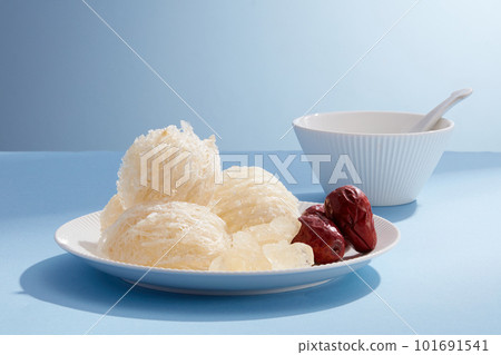 Front view of raw bird nest, rock sugar and jujube on round plate, white bowl on blue background. Advertising photo for the dish has sweet taste when cooking bird nest with jujube and rock sugar. 101691541
