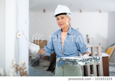 Portrait of an elderly woman painter in protective helmet who paints the walls with paint roller Portrait of an elderly woman painter in protective helmet who paints the walls with paint roller 101691834