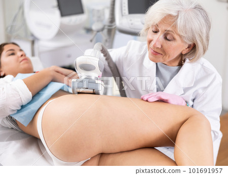 Elderly female doctor doing Rf lifting procedure on legs, buttocks and hips of young woman in beauty salon. Treatment of flabby skin Elderly female doctor doing Rf lifting procedure on legs, buttocks and hips of young woman in beauty salon. Treatment of flabby skin 101691957