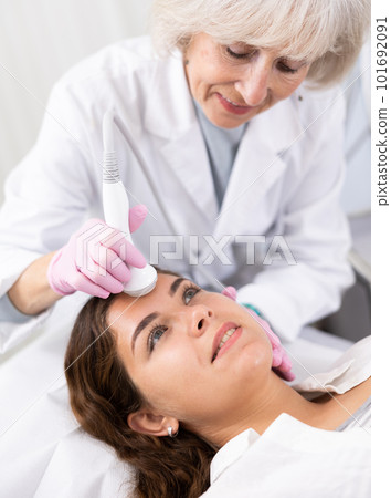 Young female visitor receiving hardware electric lifting massage with help of old female beautician in spa salon indoors Young female visitor receiving hardware electric lifting massage with help of old female beautician in spa salon indoors 101692091