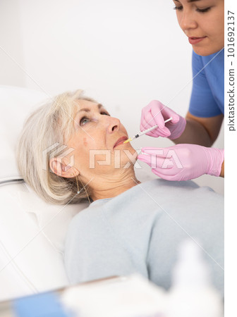 Young woman cosmetologist in uniform making injections for lip augmentation and anti-wrinkle of elderly female in esthetic clinic indoors 101692137
