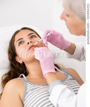 Professional mature female beautician with syringe injecting filler with hyaluronic acid in lips of young woman client during procedure in beauty salon 101692281