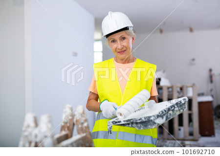 Portrait of elderly positive woman painter in protective helmet and yellow vest who paints walls with paint roller 101692710