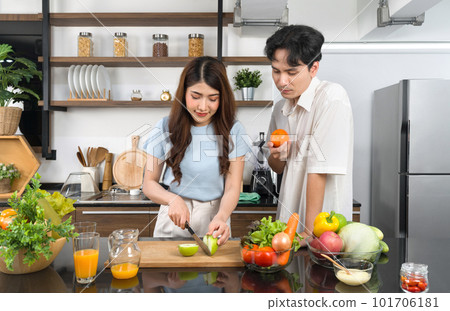 Asian couple spend time together in the kitchen. Young woman cutting green apple on a wooden chop board while her boyfriend stand beside her with an orange in his hand. Fruit juice are on the counter. Asian couple spend time together in the kitchen. Young woman cutting green apple on a wooden chop board while her boyfriend stand beside her with an orange in his hand. Fruit juice are on the counter. 101706181