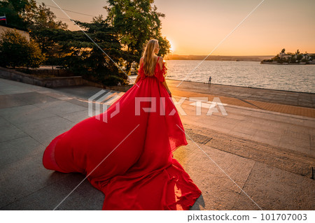 Sunrise red dress. A woman in a long red dress against the backdrop of sunrise, bright golden light of the sun's rays. The concept of femininity, harmony. 101707003