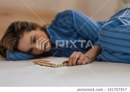 Young woman with depression lying in bed, looking at pills. 101707957
