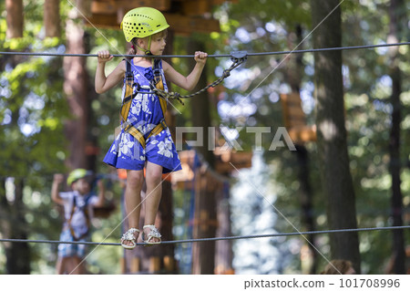 Young pretty blond child girl in safety harness and helmet on rope way on bright bokeh background. 101708996