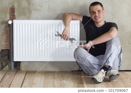 Young handsome plumber sitting on floor with wrench in hand near successfully installed heating radiator in empty room of a newly built apartment or house. Construction, maintenance and repair concept 101709067