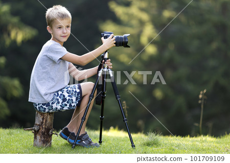 Young blond child boy sitting on tree stump on grassy clearing taking picture with tripod camera. 101709109