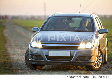 Young attractive long-haired woman inside shiny silver car at steering wheel on gravel empty road on bright blurred summer sunny green field and misty sky background. Young attractive long-haired woman inside shiny silver car at steering wheel on gravel empty road on bright blurred summer sunny green field and misty sky background. 101709115